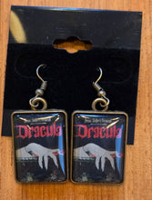 Load image into Gallery viewer, A pair of rectangular gothic-style earrings featuring a Dracula-themed design, displayed on a black backing card next to a yellow tape measure on a wooden surface.